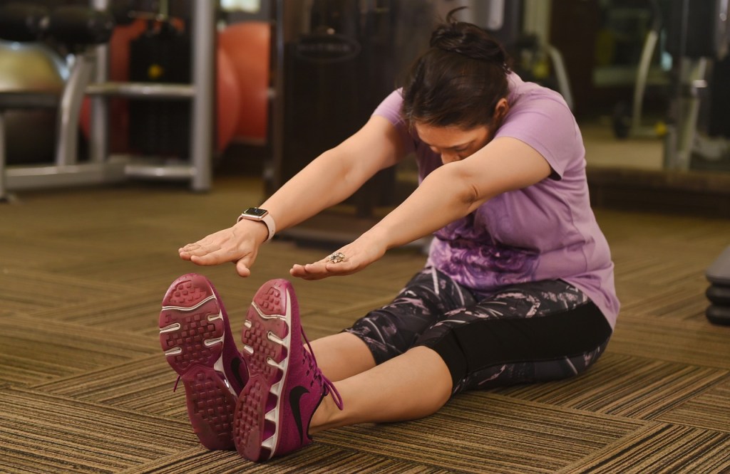 Woman in a gym sitting on the floor stretching forward to touch her toes.