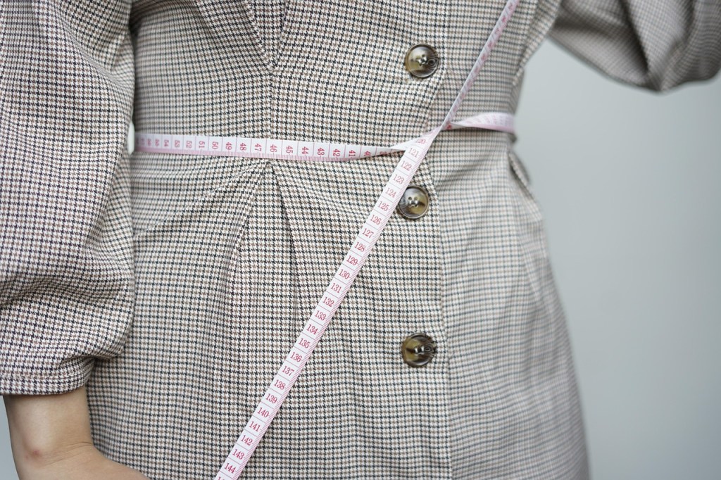 A person wearing a beige dress measuring their waist with a tape measure, focusing on fitness and body measurement tracking.