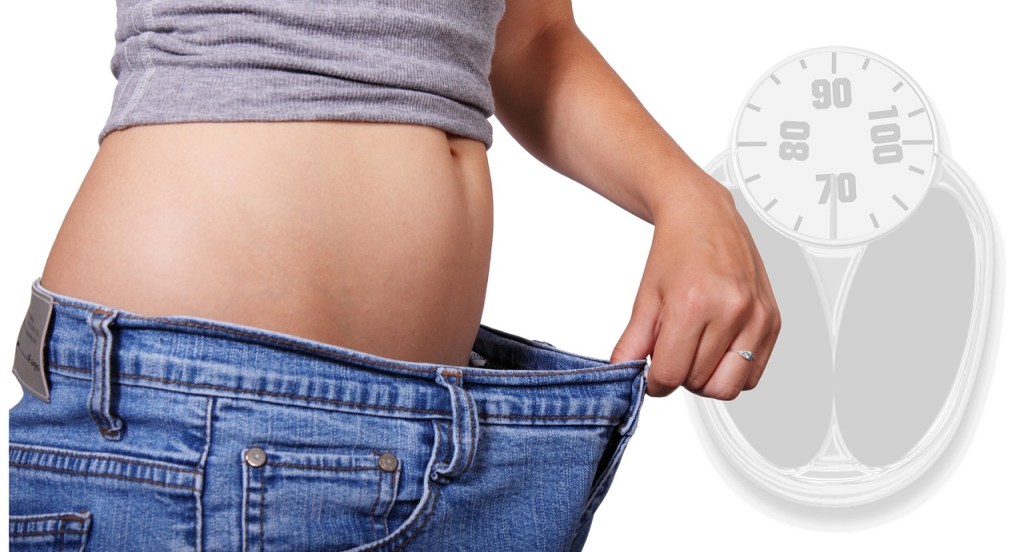 Person pulling loose jeans' waistband, indicating weight loss, with a faint weighing scale in the background.